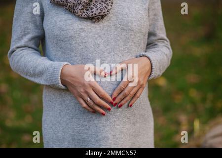 Schwangerschaft, Liebe, Familie und Menschen Konzept - Nahaufnahme einer schwangeren Frau, die eine Hand-Herz-Geste auf dem Bauch macht Stockfoto