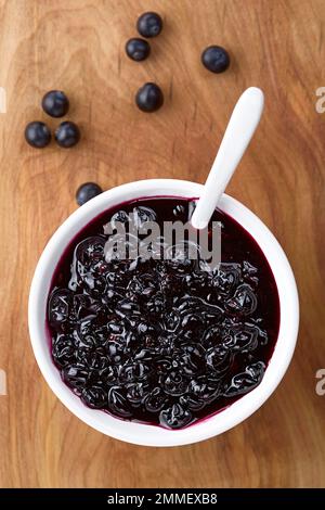 Frische hausgemachte Marmelade aus patagonischen Calafatbeeren (lat. Berberis heterophylla) serviert in einer weißen Schüssel mit Löffel, über Kopf fotografiert auf Holz Stockfoto