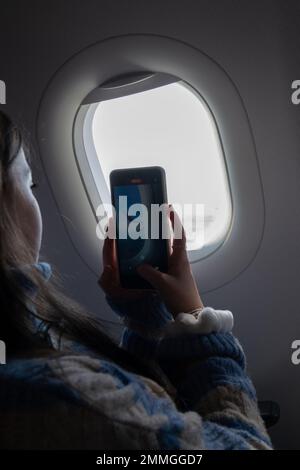 Ein junges Mädchen, das im Flugzeug am Fenster sitzt und beim Fliegen am Himmel Fotos macht. Kinder, die mit Flugzeugen reisen. Teenager Passagiere. Stockfoto
