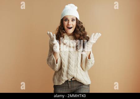 Hallo Winter. Porträt einer überraschend stilvollen 40-jährigen Frau in beigefarbenem Pullover, Fäustlingen und Hut isoliert auf beigefarbenem Hintergrund. Stockfoto