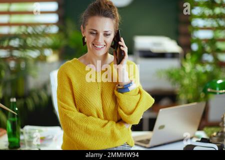 Lächelnde Eigentümerin eines kleinen Unternehmens mittleren Alters in einem gelben Pullover im modernen grünen Büro, die auf einem Smartphone spricht. Stockfoto