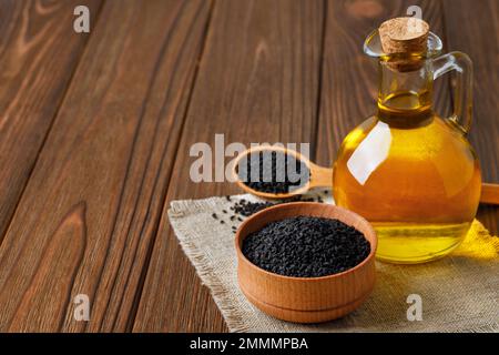 Schwarzes Kreuzkümmelöl in Glasflasche und Samen in Schüssel und Löffel auf dem Tisch Stockfoto