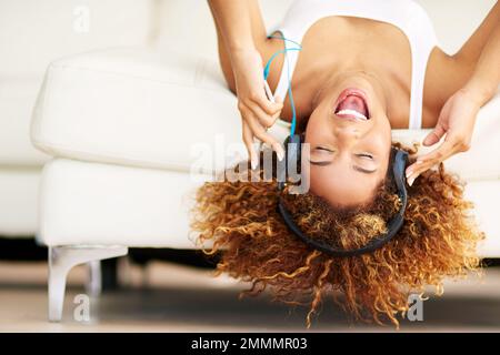 Ich kann nicht einen Tag ohne Musik auskommen. Eine junge Frau, die Musik hört, während sie kopfüber auf ihrer Couch liegt. Stockfoto