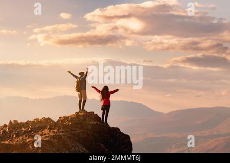 Zwei glückliche Wanderer stehen mit offenen Armen am Aussichtspunkt bei Sonnenuntergang hoch in den Bergen Stockfoto