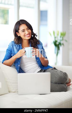 Es geht nichts über eine frisch gebrühte Tasse Kaffee. Eine junge Frau, die zu Hause auf ihrem Laptop im Internet surft. Stockfoto