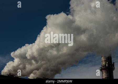 Weißer und grauer Rauch und Dampf aus einem hohen Betonkamin am hellblauen Himmel. Stockfoto