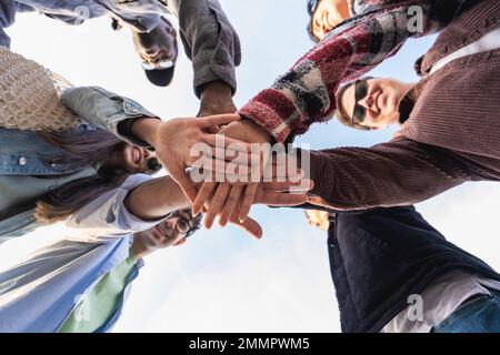 Ein Blick von unten auf eine multiethnische Gruppe junger Freunde, die im Kreis stehen und sich in Richtung der Mitte die Hände zusammenschließen Stockfoto