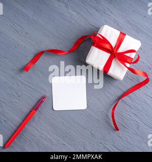 Stilvolle, minimalistische Postkarte zum Valentinstag. Geschenk in weißem Papier mit rotem Band, leerem weißem Blatt und rotem Stift auf grauem 2021-Trendhintergrund, Stockfoto