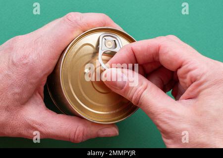 Die Hand eines Erwachsenen hält eine Dose Dosenfutter, die zweite Hand zieht den Schlüssel und öffnet die Dose. Draufsicht auf grünem Hintergrund mit Kopierbereich. Nahaufnahme Stockfoto