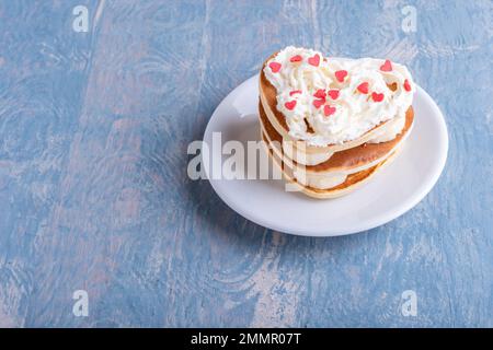 Romantisches Frühstück im Bett. Hausgemachter Pfannkuchen in Herzform. Pfannkuchen in einem Stapel mit Banane und mit weißer Creme und Rot dekoriert Stockfoto