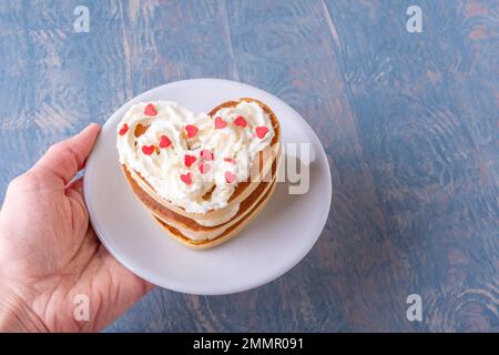 Kreatives Frühstück am Valentinstag. Weibliche Hand hält einen weißen Teller mit einem Stapel hausgemachter herzförmiger Pfannkuchen, dekoriert mit weißer Creme und Stockfoto
