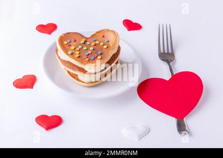 Hausgemachte herzförmige Pfannkuchen, dekoriert mit Zuckerbonbons auf einem weißen Teller, einer Gabel und einer roten Herzform auf weißem Hintergrund mit Herzen, Draufsicht, Stockfoto