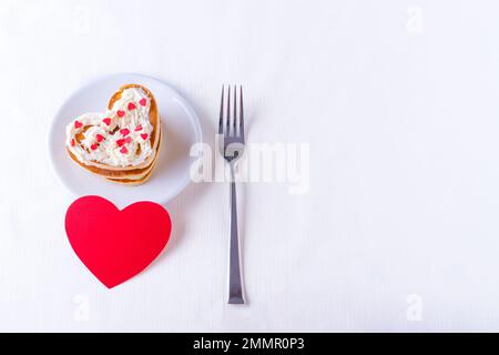 Hausgemachte herzförmige Pfannkuchen, dekoriert mit Sahne- und Zuckerbonbons auf einem weißen Teller, Gabel und rotes, rotes, leeres Herz auf weißem Hintergrund, Draufsicht, Stockfoto