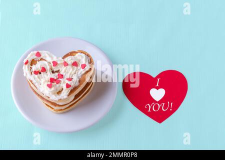 Kreatives Frühstück am Valentinstag. Hausgemachter herzförmiger Pfannkuchen mit weißer Creme und roten Herzen auf einem weißen Teller auf blauem Hintergrund Stockfoto