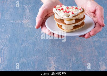Kreatives Frühstück im Bett. Weibliche Hand hält einen weißen Teller mit einem Stapel hausgemachter herzförmiger Pfannkuchen, dekoriert mit weißer Creme und roten Herzen Stockfoto