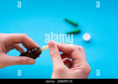 Die Hand einer Frau mit einer Pipette sammelt Flüssigkeit aus einer dunkelbraunen Flasche auf blauem Hintergrund. Kosmetisches Hautpflegekonzept. Medizinisches Konzept Stockfoto