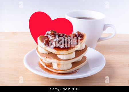 Kreatives Frühstück zum Valentinstag, Muttertag, Geburtstag. Hausgemachte herzförmige Pfannkuchen mit Beerenmarmelade und Bananen auf weißem Teller, Gabel, Becher Stockfoto