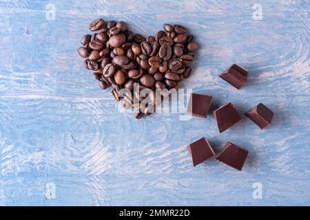 Symbol der Liebe zu Kaffee und Schokolade. Herzform aus Kaffeebohnen und Stücken schwarzer bitterer Schokolade auf blauem Holzhintergrund, Nahaufnahme Stockfoto