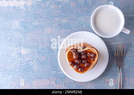 Hausgemachte herzförmige Pfannkuchen mit Beerenmarmelade, weißer Milchbecher und Gabel auf blauem Holztisch, Kopierbereich, Draufsicht Stockfoto