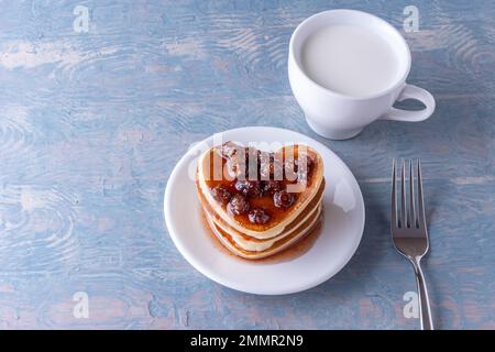 Süßes hausgemachtes Frühstück für Kinder. Hausgemachte herzförmige Pfannkuchen mit Beerenmarmelade, weißer Milchbecher und Gabel auf blauem Holztisch, Kopierbereich, Top vi Stockfoto