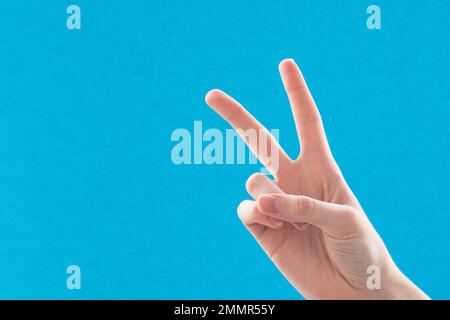 Nahaufnahme einer weiblichen Hand mit zwei Fingern, ein Symbol des Friedens oder des Sieges. Unterschreiben Sie für den Buchstaben V in Gebärdensprache. Auf blauem Hintergrund Stockfoto