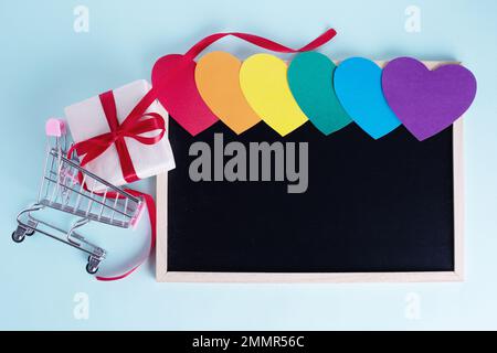 Geschenkbox in Einkaufswagen, bunt regenbogenfarbene Papierherzen und eine leere Tafel mit Kopierbereich auf hellblauem Hintergrund. Saisonangebot, DIS Stockfoto