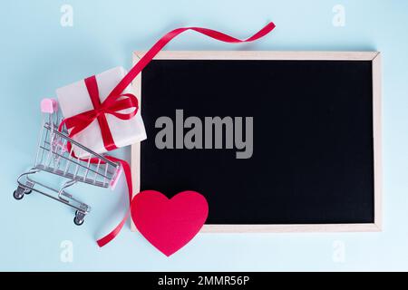 Geschenkbox in Einkaufswagen mit rotem Papierherz und leerer Tafel mit Kopierbereich auf blauem Hintergrund. Saisonangebot, Rabatte, Black friday-Konzert Stockfoto