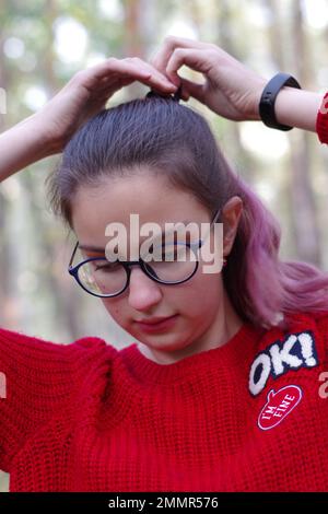 Porträt einer süßen Weißen in blauer Brille, rotem Pullover, verknoteten Haaren im Wald. Stockfoto