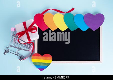 Geschenkbox in Einkaufswagen, bunt regenbogenfarbene Papierherzen und eine leere Tafel mit Kopierbereich auf hellblauem Hintergrund. Saisonangebot, DIS Stockfoto