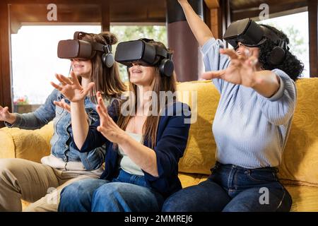 Drei Freundinnen, die auf einer Couch sitzen und 3D VR-Headsets tragen, machen Handgesten, während sie eine virtuelle Welt im Metaversum erkunden. Stockfoto