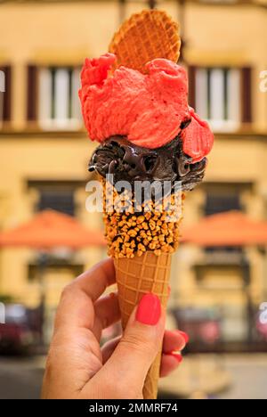 Halten Sie ein handgemachtes Schokoladen- und Himbeergelato in der Hand, und genießen Sie den Blick auf verschwommene Häuser und Lichter auf einer Fußgängerstraße im Centro Storico, Florenz, Italien Stockfoto
