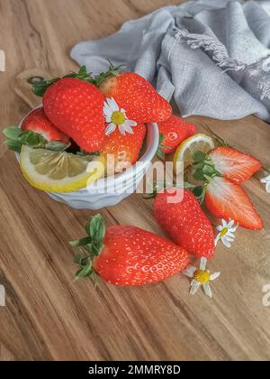 Blick auf ganze und halbierte Erdbeeren in einer weißen Schüssel und auf einem Holztisch, dekoriert mit Zitronenscheiben und Blütenblättern, dekorative Fabrik Stockfoto