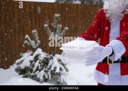 Lebensmittelliefercontainer in den Händen des Weihnachtsmanns im Freien im Schnee. Weihnachtsabend-Angebot. Gebrauchsfertige heiße Bestellung, Einweg-Kunststoffbox. Neues y Stockfoto