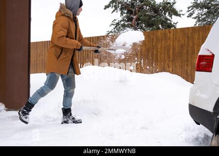 Ein Mann im Winter reinigt Schnee mit einer Schaufel im Hof eines Hauses auf dem Parkplatz. Schneefall, schwierige Wetterbedingungen, das Auto steht still, digg Stockfoto