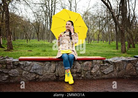 Glückliche Seniorin in gelbem Regenmantel mit gelbem Regenschirm spricht mit einem Smartphone und läuft im Park. Mit entfernten Lieben in Kontakt bleiben Stockfoto