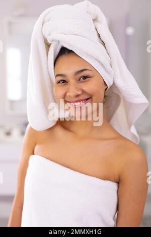 Vertikales Porträt einer glücklichen, birassischen Frau, die im Badezimmer ein lächelndes Handtuch trägt, mit Kopierbereich. Entspannung, Selbstversorgung, Schönheit, Freizeit und Lifestyle con Stockfoto