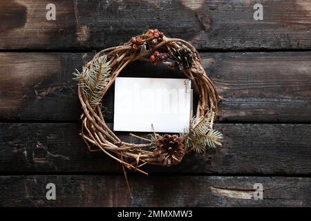 Neujahrskranz und in der Mitte eine weiße Papierkarte für Text auf einem alten Holztisch, Dekor für die Feiertage, ein Kranz von einer trockenen Weinrebe, eine pla Stockfoto