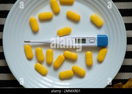 Ein elektronisches Thermometer liegt neben gelben Pillen auf einem Teller auf dem Tisch in der Küche, Medizin und Apotheke, Gesundheit und Behandlung, Pillen und Ameise Stockfoto