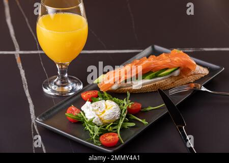 Brxchetta mit Avocado und gesalzenem Lachs. Auf einem Teller mit pochiertem Ei, im Rahmen eines Glases mit Orangensaft und einem Teller mit Bruscheta. Schwarz Stockfoto