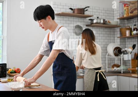 Fokussierter junger asiatischer Mann in der Schürze knettet rohen Teig mit einer Rolle, macht Teig, während seine Freundin das Geschirr in der Küche wäscht. Stockfoto