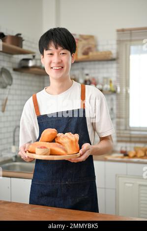 Ein glücklicher und gutaussehender junger asiatischer Mann mit Schürze hält ein Tablett mit frischem Gebäck in seiner Küche. Backen zu Hause Konzept Stockfoto