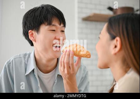 Fröhlicher und verspielter junger asiatischer Freund, der seiner Freundin in der Küche ein Croissant fütterte und Spaß beim Frühstück in der Küche hatte. Stockfoto