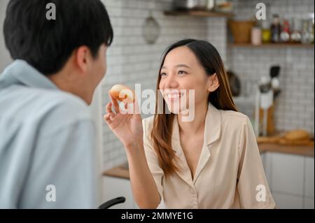 Hübsche und reizende junge Asiatin, die ihrem Freund Donuts füttert, und sich in der Küche amüsiert. Stockfoto