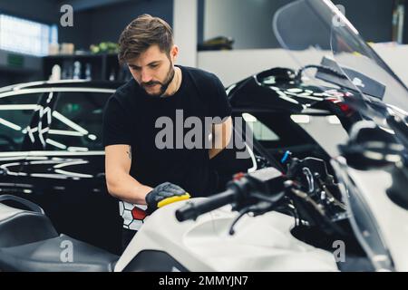 Fokussierter Mechaniker, der die Oberfläche eines weißen Motorrads in seiner Garage poliert. Schwarzes Auto im Hintergrund. Mittlere Indoor-Aufnahme. Speicherplatz kopieren. Hochwertiges Foto Stockfoto