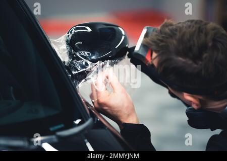 PPF-Wickelvorgang. Rückansicht eines nicht erkennbaren professionellen Mechanikers beim Auftragen von Schutzfolie auf den Seitenspiegel. Hochwertiges Foto Stockfoto