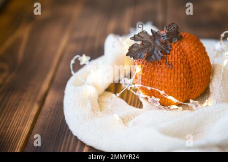 Orangefarbener Kürbis, eingewickelt in einem warmweißen Karo mit Lichtern von Girlanden, auf einem Holztisch mit Kopierraum. Herbststimmung, halloween, Ernte, Thanksgiving, Stockfoto