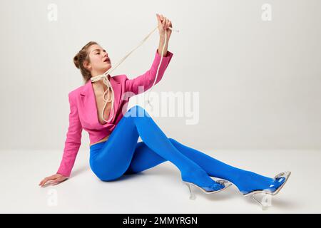 Emotionales junges, stilvolles Mädchen in blauen Strumpfhosen und rosa Croptop posieren, auf dem Boden isoliert auf grauem Hintergrund seltsame Posen machen. Das Konzept von "seltsam" Stockfoto