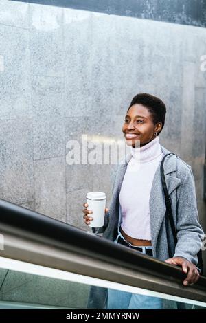 Hochwinkel einer optimistischen afroamerikanischen Frau in stilvoller Oberbekleidung. Sie hält eine Tasse Kaffee ohne Abfall und sie lächelt und fährt tagsüber in der Stadt Treppen aus der Untergrundbahn Stockfoto
