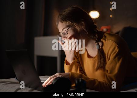 Müde junge Frau, gähnt, auf dem Bett mit einem Laptop liegt, faule Studentin, die Hausaufgaben macht, schläfriges Mädchen, das im Schlafsaal am Computer arbeitet, schlaflose Nacht, Mangel Stockfoto