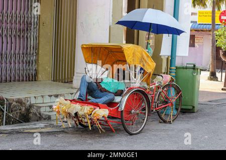 Georgetown, Penang, Malaysia - November 2012: Ein Rikscha-Fahrer macht ein Nickerchen in seinem Fahrzeug, das auf einer Straße geparkt ist. Stockfoto
